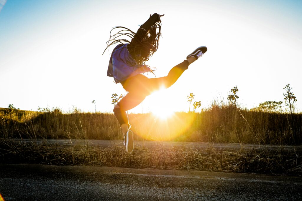 The image is a black-and-white negative photograph of a dancer performing a leap outdoors in a field. The dancer is captured mid-motion wherein the back leg is extended high in the air and the other is bent lower to the ground. The dancer's long braids flows upwards, capturing the energy of this mid-motion leap